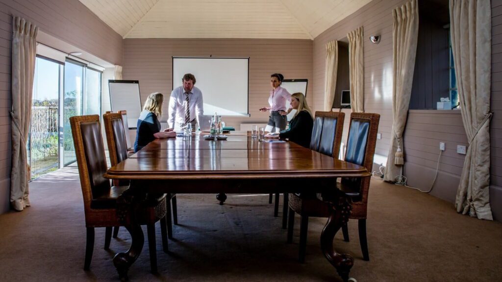 Four people are having a meeting in a conference room with a large wooden table, flip chart, and projection screen.