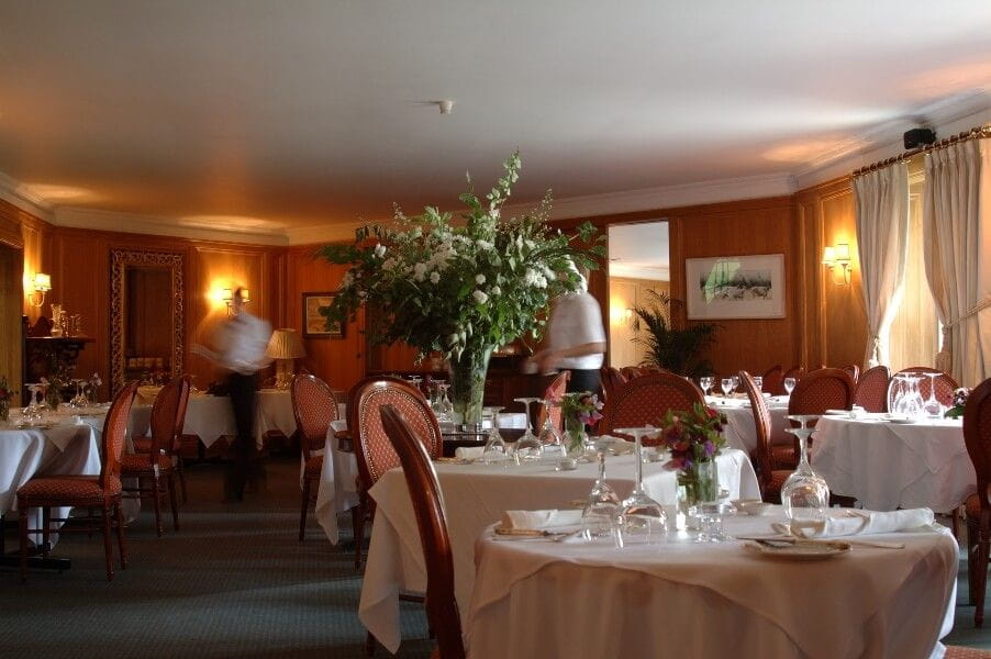 Elegant restaurant dining room with round tables set with white tablecloths, glassware, and flowers. Two blurred waiters move around a large central floral arrangement.