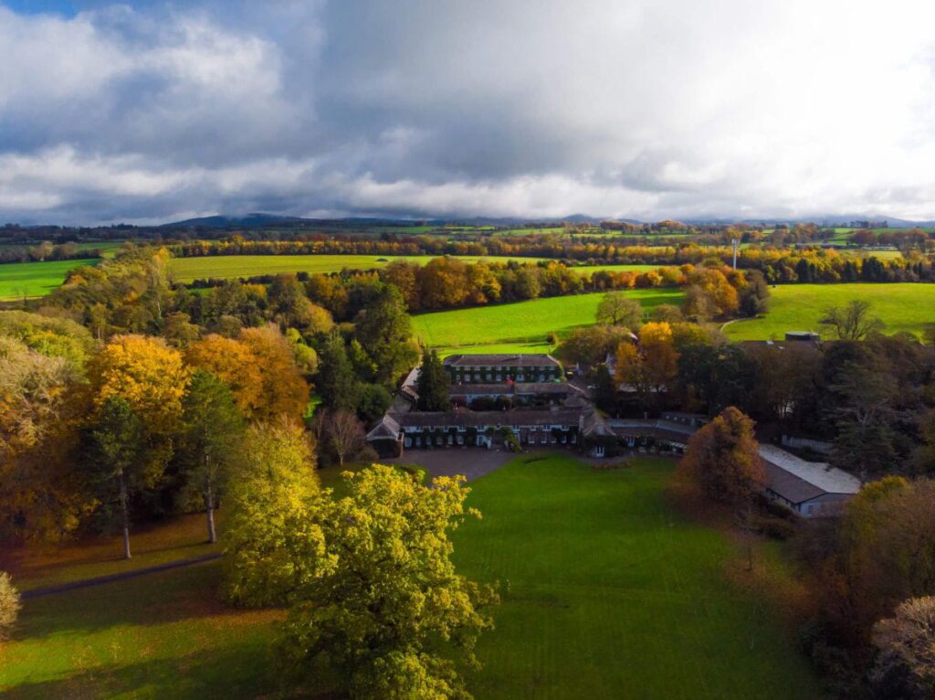 Aerial view of a countryside estate surrounded by green fields, autumn trees, and distant hills under a cloudy sky.