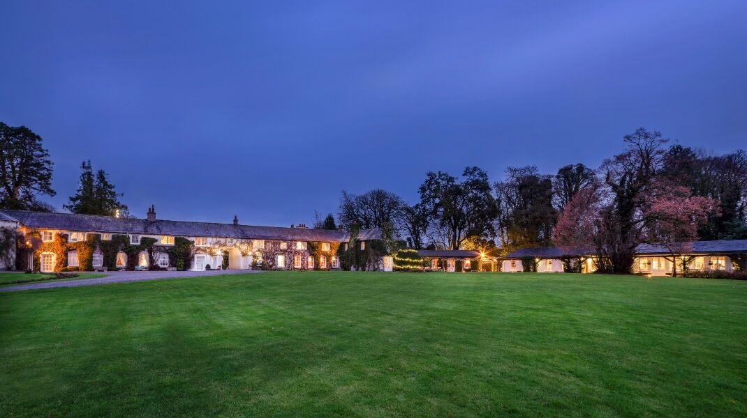 Large historic stone building with lit windows sits on a wide, neatly mown lawn, surrounded by tall trees under a dusky blue sky.