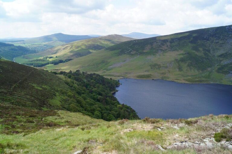 A view of a dark blue lake surrounded by green hills and scattered trees under a partly cloudy sky.