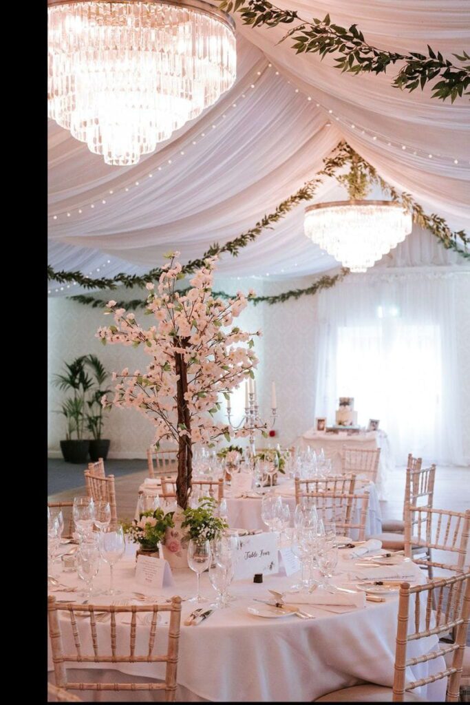 Elegant banquet hall with draped white fabric ceiling, chandeliers, round tables set with glassware, floral centrepieces, and wooden chairs. Greenery decorates the ceiling.