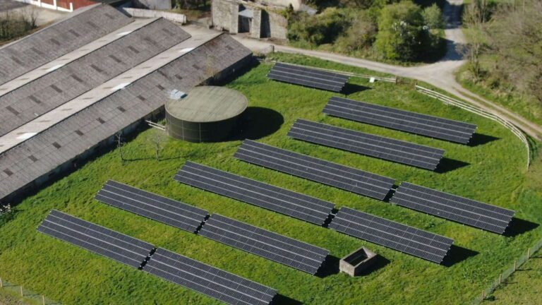 Rows of solar panels are installed on a grassy field next to a large building and a circular water tank, viewed from above.