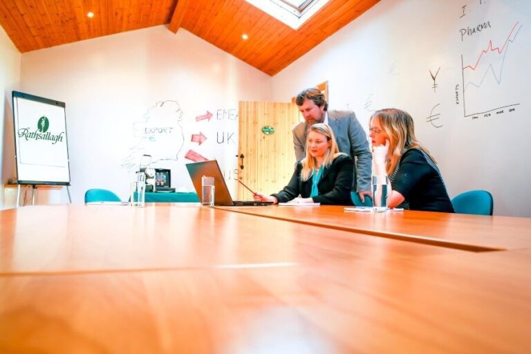 Three people in business attire sit and stand around a laptop in a meeting room, with charts and diagrams on the wall and a flipchart in the background.