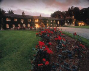 A large, ivy-covered country house with glowing windows stands behind a flowerbed of red roses and a gravel drive at dusk.