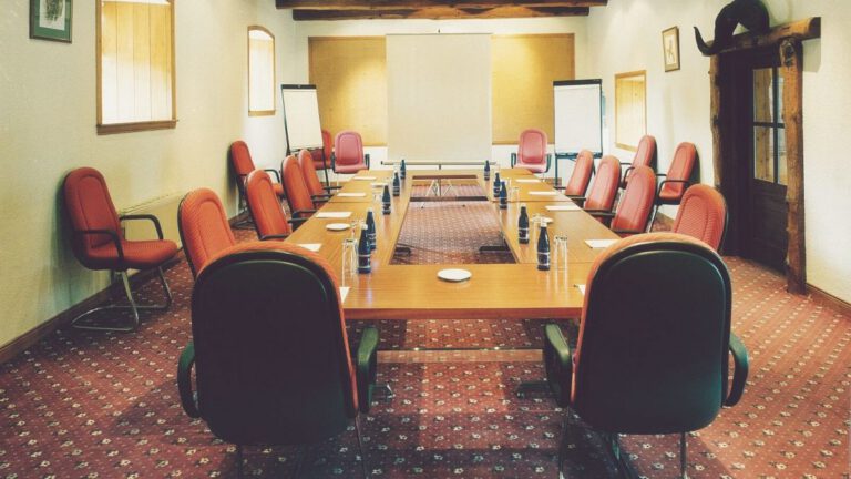 A conference room with a U-shaped table arrangement, red chairs, water bottles, notepads, flipcharts, and a carpeted floor. The room has beige walls and wood accents.
